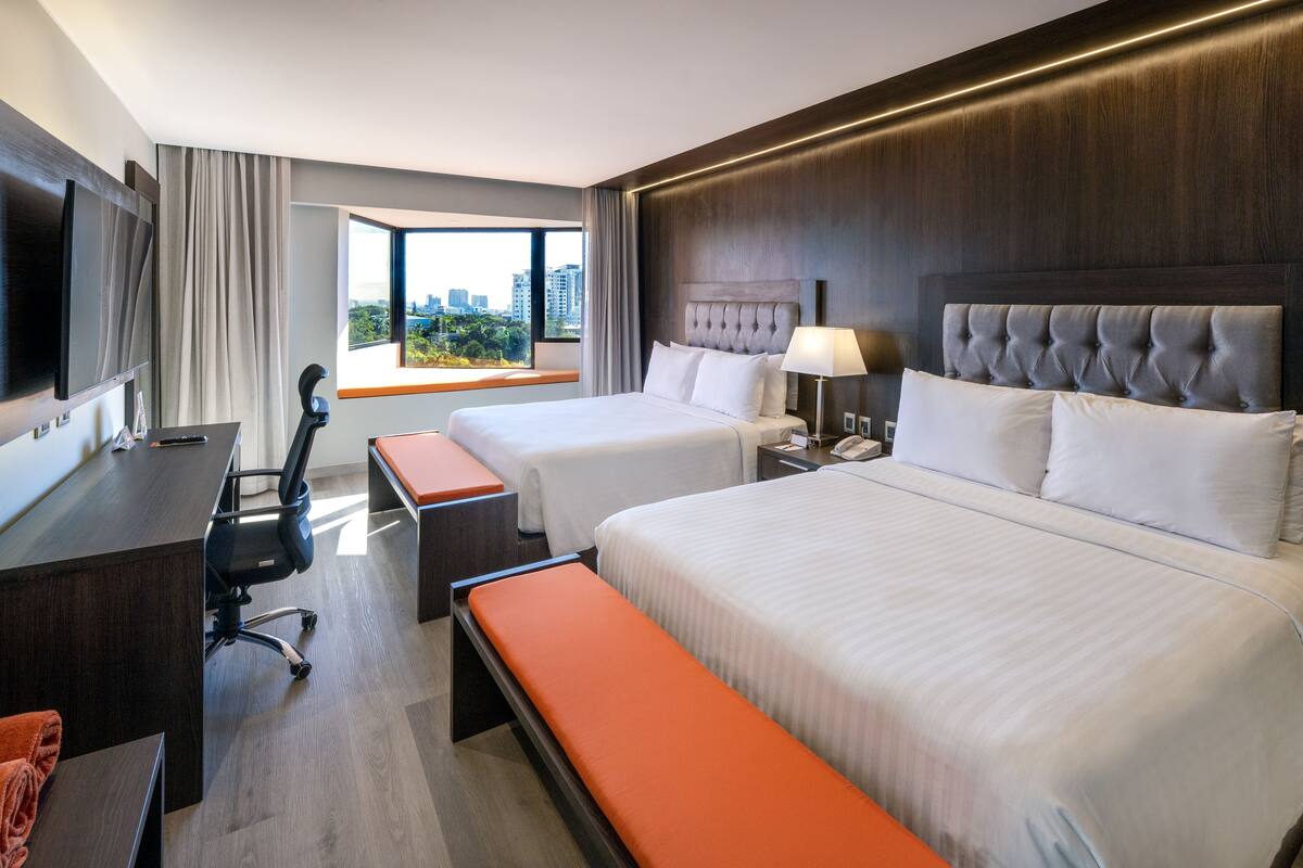 A hotel room with two beds, a desk, a chair, a TV, and a window with a city view.