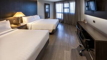 Habitación de hotel con dos camas, un escritorio con silla, televisor montado en la pared y una ventana con cortinas.