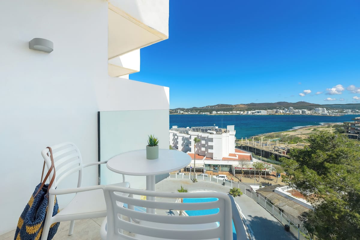 A balcony with a white table and chairs, a small potted plant, and a view of the sea and buildings in the distance.