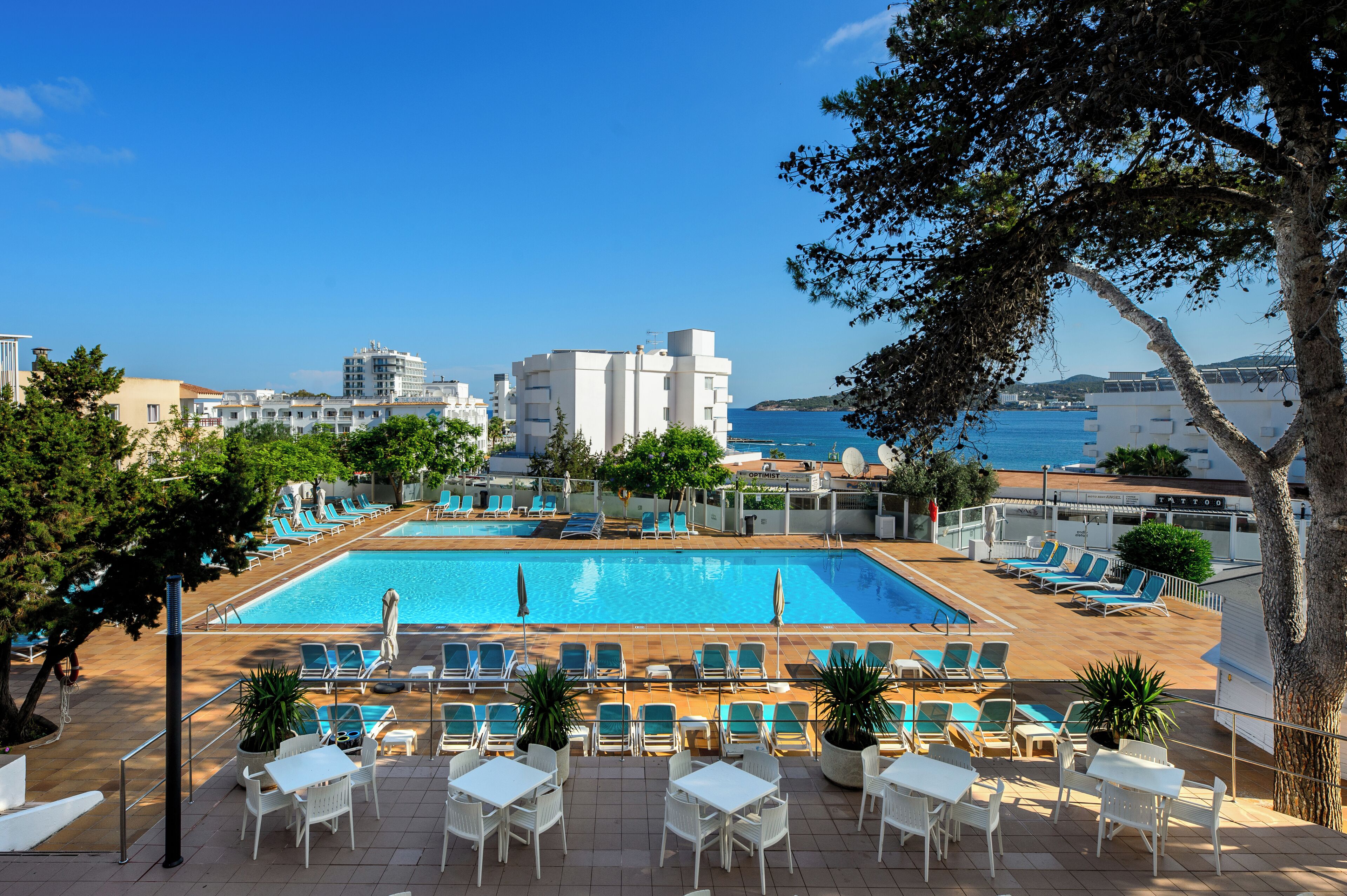 A pool area with lounge chairs and tables, a clear view of the sea, and buildings in the background.