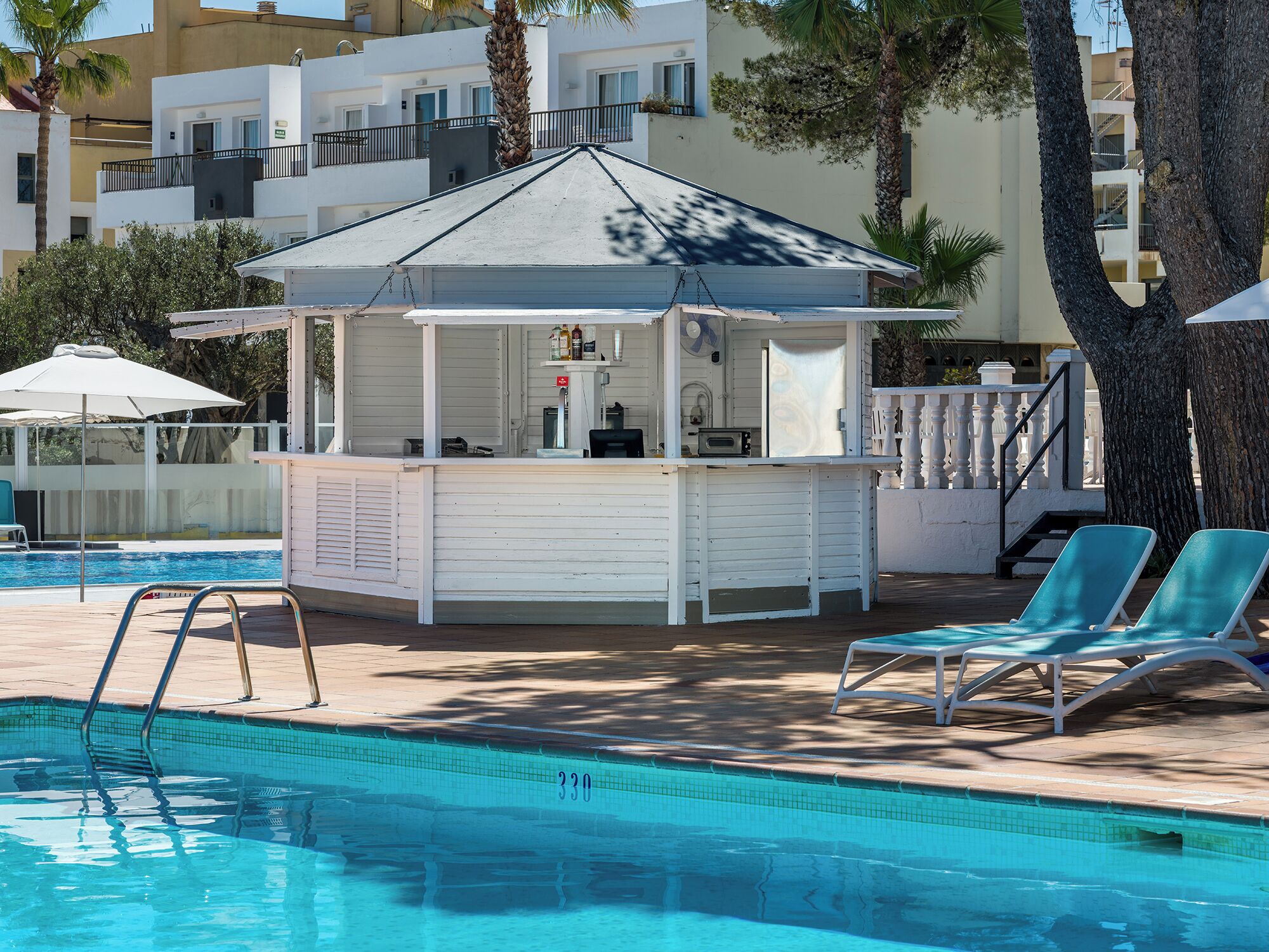 Eine Poolbar mit weißer Fassade, ein Pool mit klarem, blauem Wasser und zwei Liegestühle.