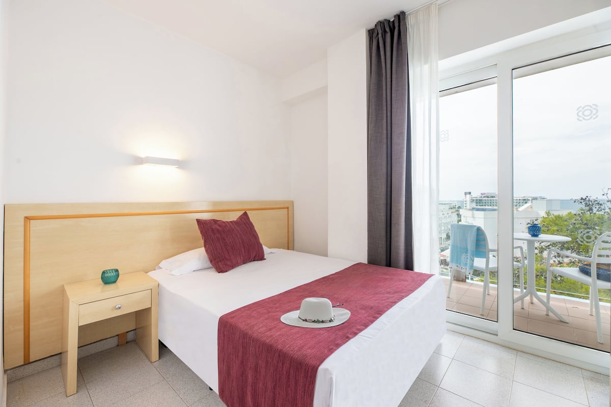 A hotel room with a bed, a nightstand, a window with a view of the city, and a balcony with outdoor furniture.