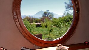 A bedroom with a round window offering a view of a mountain and greenery. There are two pillows and a blanket on the bed.