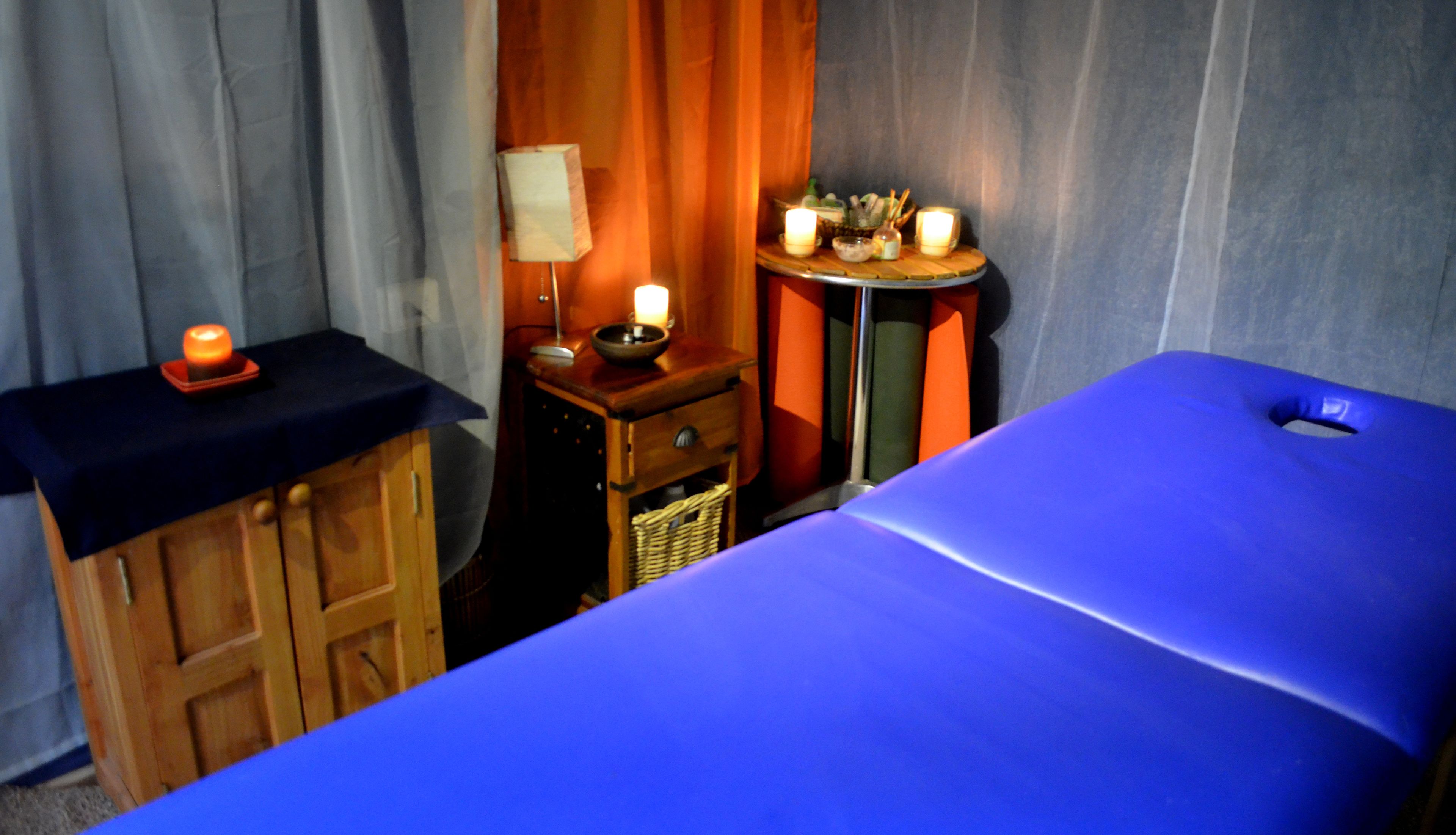 Ein Raum mit einem blauen Massagetisch, einem Holzschrank und einem kleinen Tisch mit Kerzen und einer Lampe.