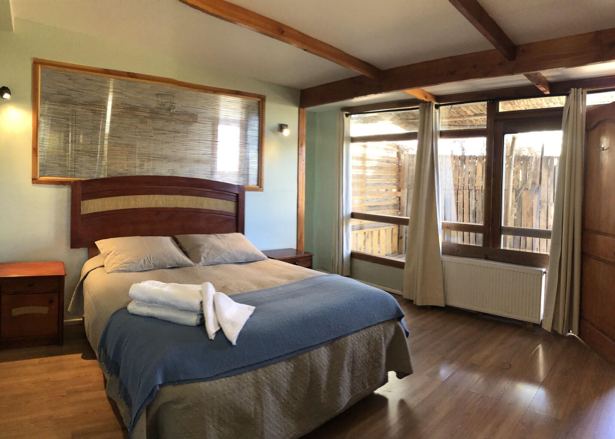 Une chambre à coucher avec une tête de lit en bois, un grand lit recouvert d’une couverture bleue et beige, une table de chevet et une fenêtre avec des stores en bois.