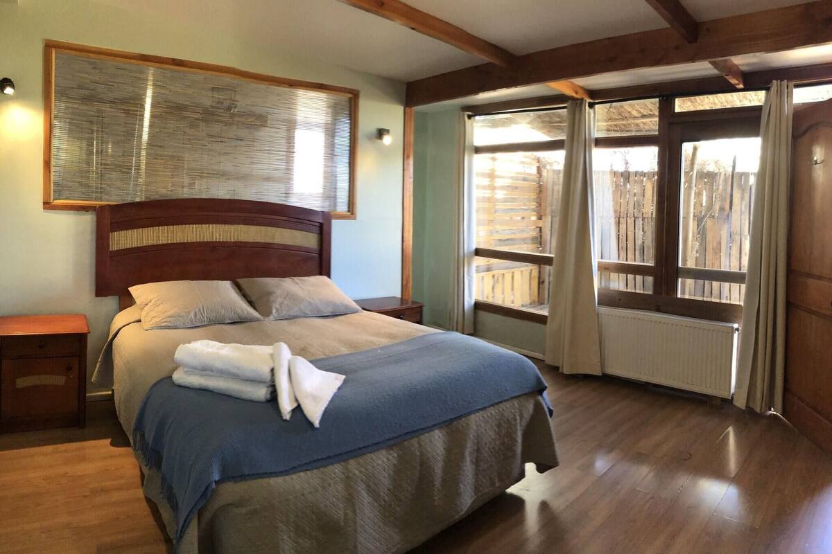 A bedroom with a wooden headboard, a large bed with a blue and beige bedspread, a nightstand, and a window with wooden blinds.