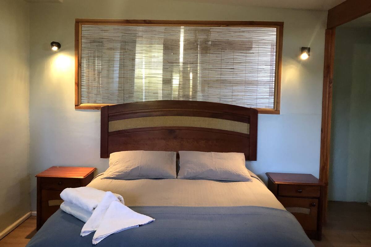 A neatly made bed with a wooden headboard, two bedside tables, and a window with blinds.