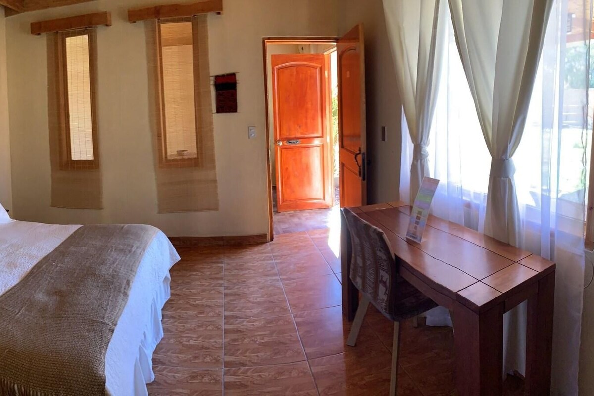 Un dormitorio con cama, mesita de noche, escritorio de madera y ventana con cortinas.