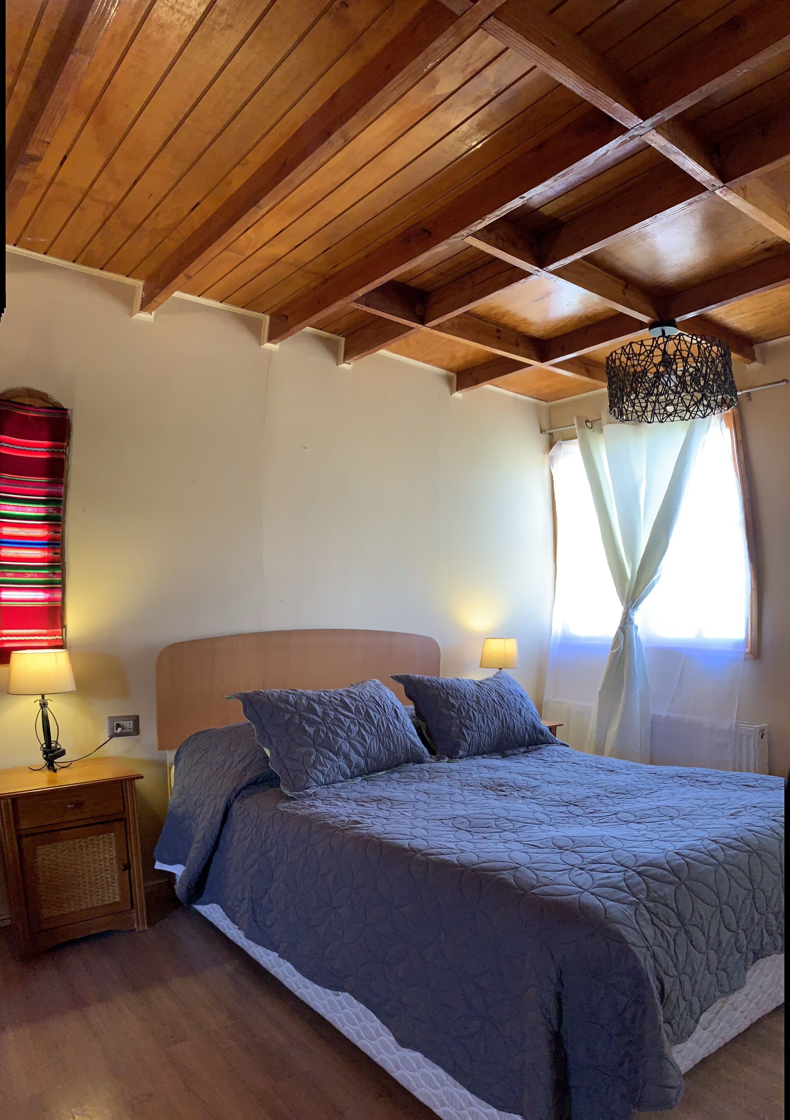 A bedroom with a wooden ceiling, a bed with a grey quilt, two bedside lamps, and a window with sheer curtains.