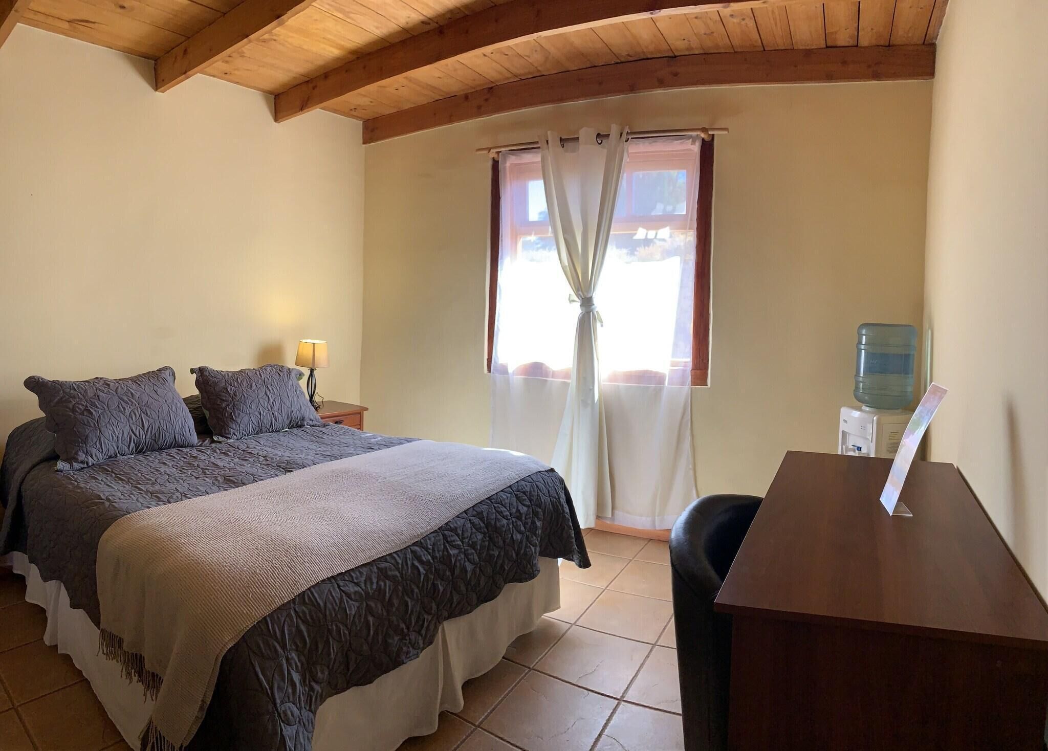 Une chambre à coucher comprenant un lit, une table de chevet, une lampe, une fenêtre avec des rideaux et un distributeur d’eau.