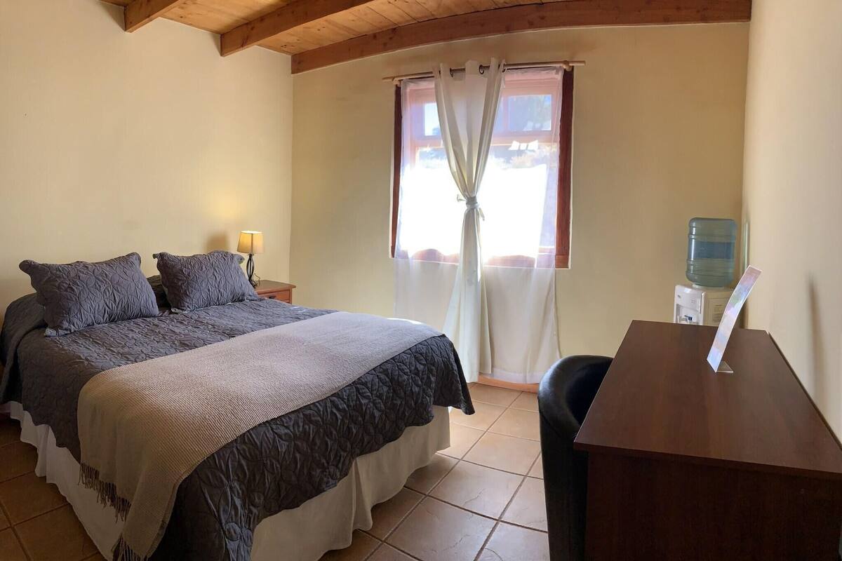 A bedroom with a bed, bedside table, lamp, window with curtains, and a water dispenser.