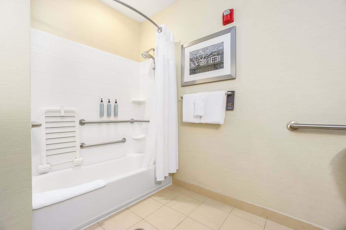 A bathroom with a white bathtub, handrails, a shower curtain, a framed picture, and a towel rack.