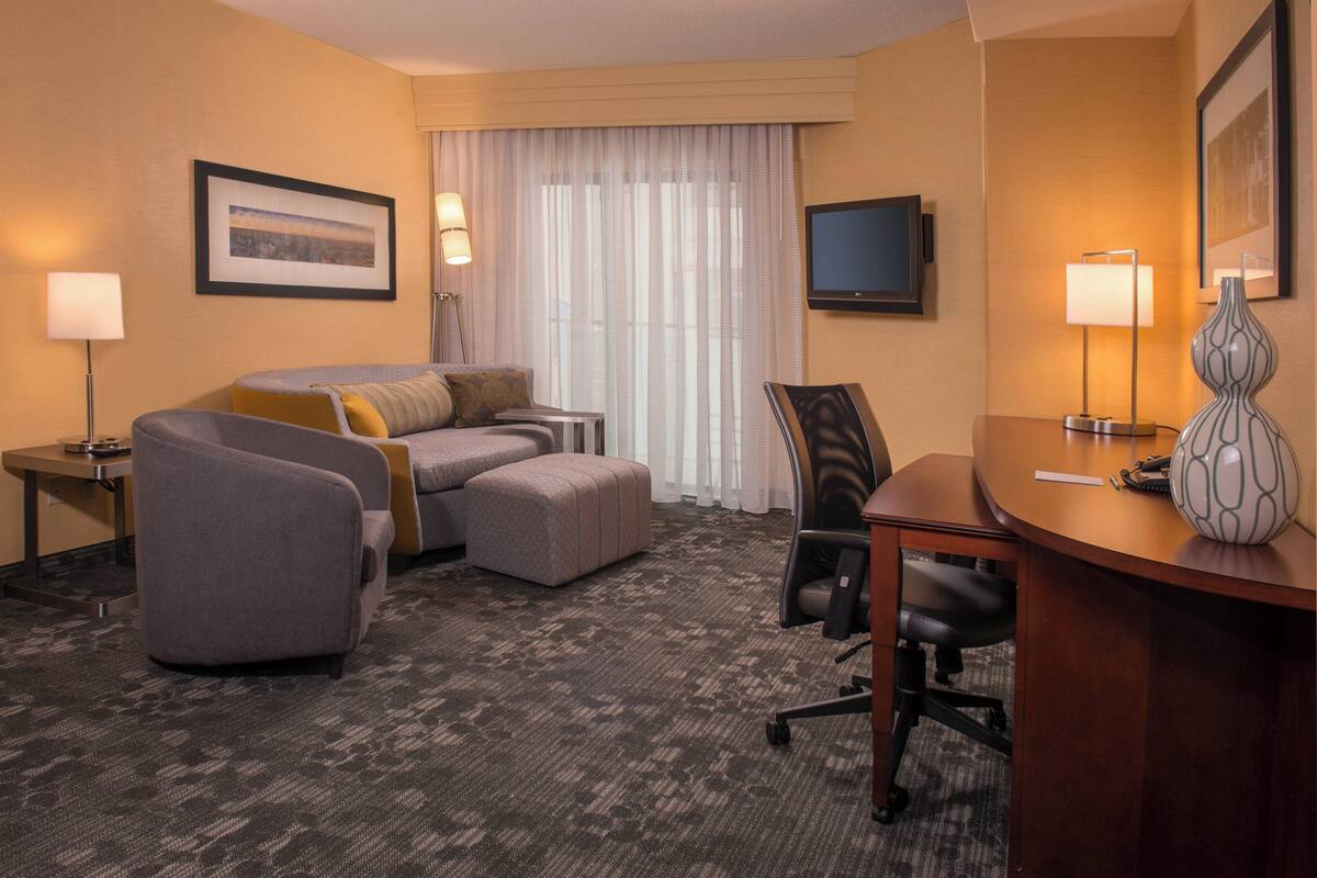 A hotel room with a sofa, ottoman, desk, chair, lamp, TV, and framed picture on the wall.