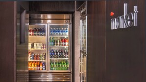 Un refrigerador comercial repleto de bebidas y aperitivos, ubicado en un cuarto con revestimiento de madera oscura y un letrero que dice 'market'.