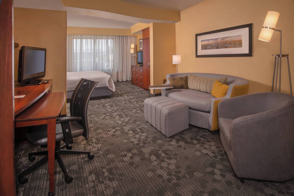 A hotel room with a desk, two beds, a sofa, a lamp, and a framed picture on the wall.