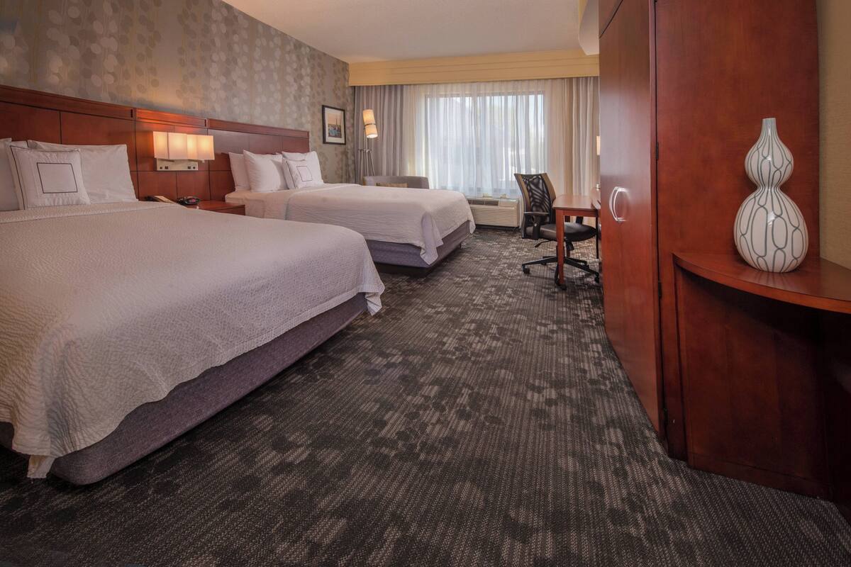 A hotel room with two beds, a desk, a chair, a lamp, and a vase.