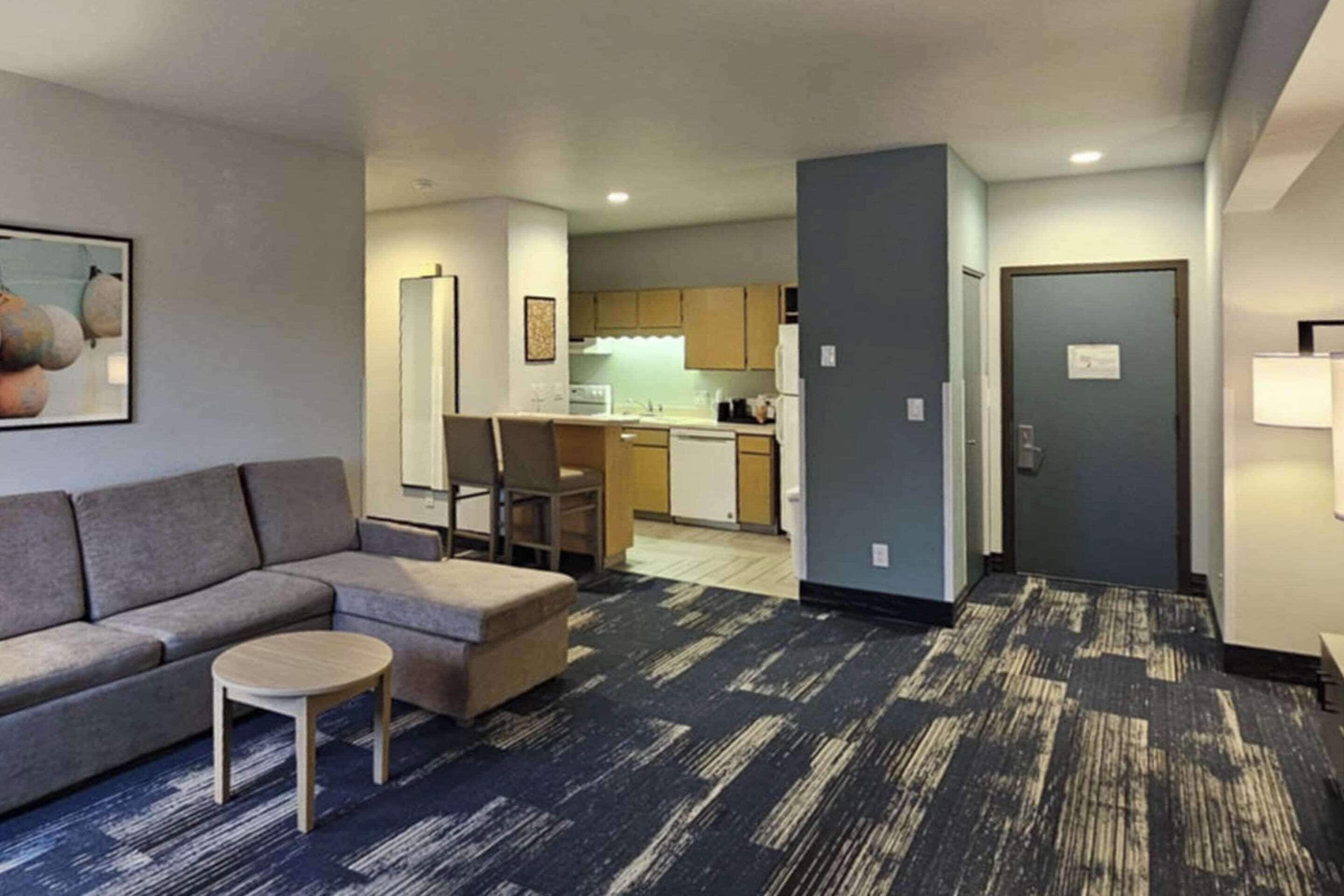 A modern hotel room with a sofa, ottoman, and a small table. A kitchen area is visible in the background.