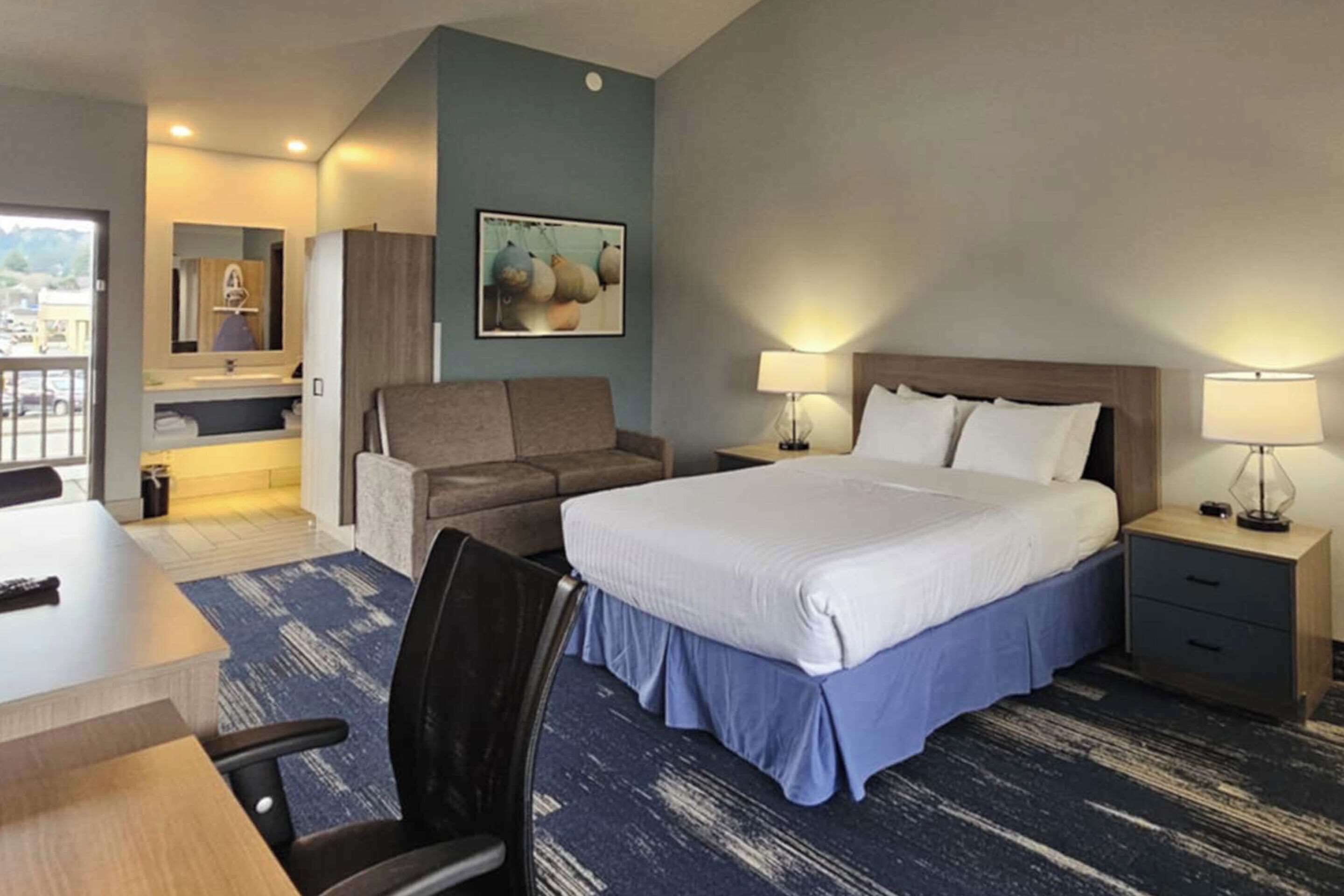 A hotel room with a large bed, a desk, a chair, a small couch, a nightstand, and a bathroom with a sink and mirror.