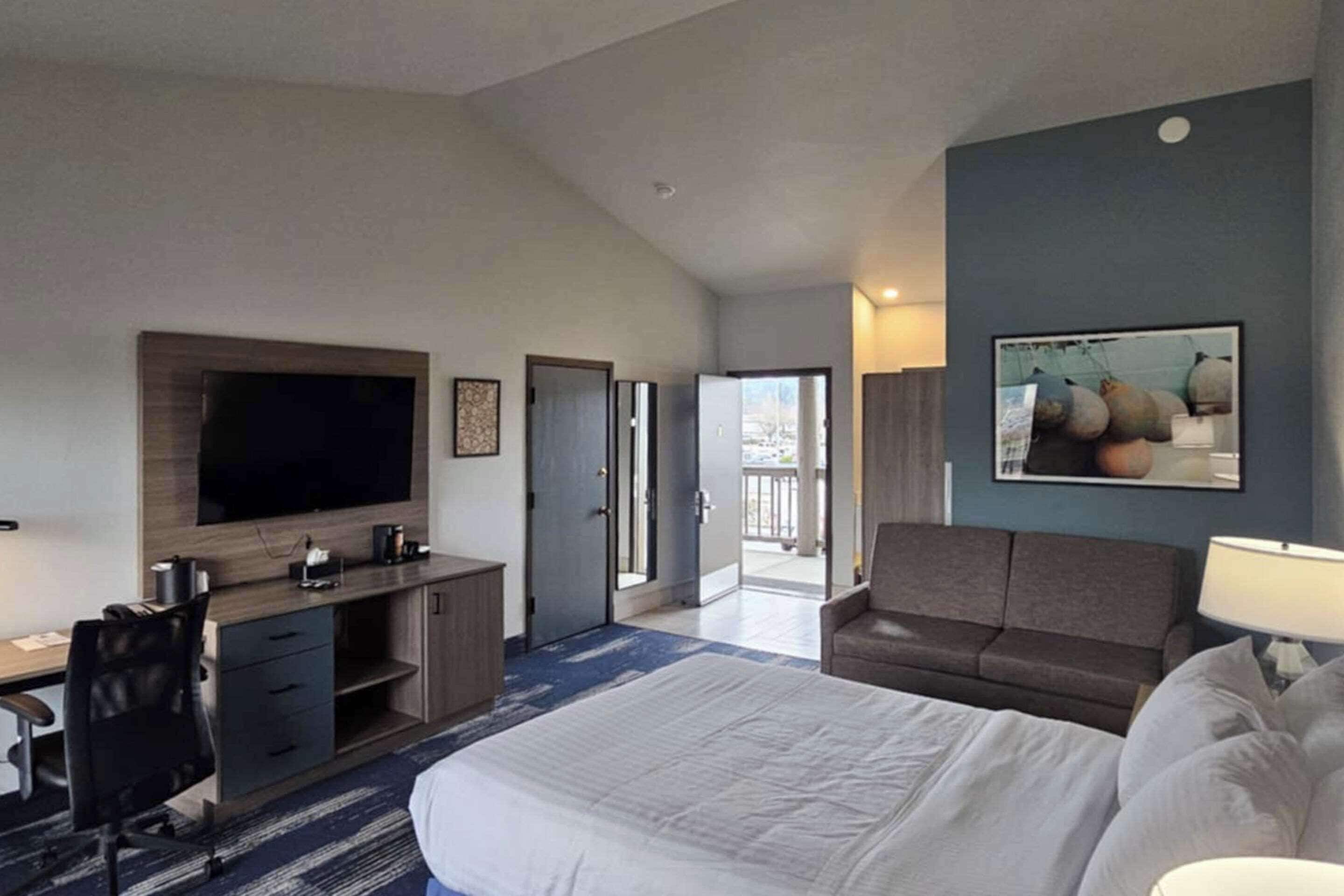 Habitación de hotel con una cama grande, un escritorio con televisor, un sofá y una puerta que da a un balcón.