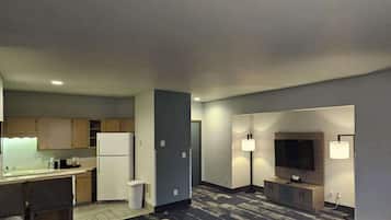 A compact hotel room with a kitchenette, a seating area with a sofa and ottoman, a flat-screen TV, and a lamp.