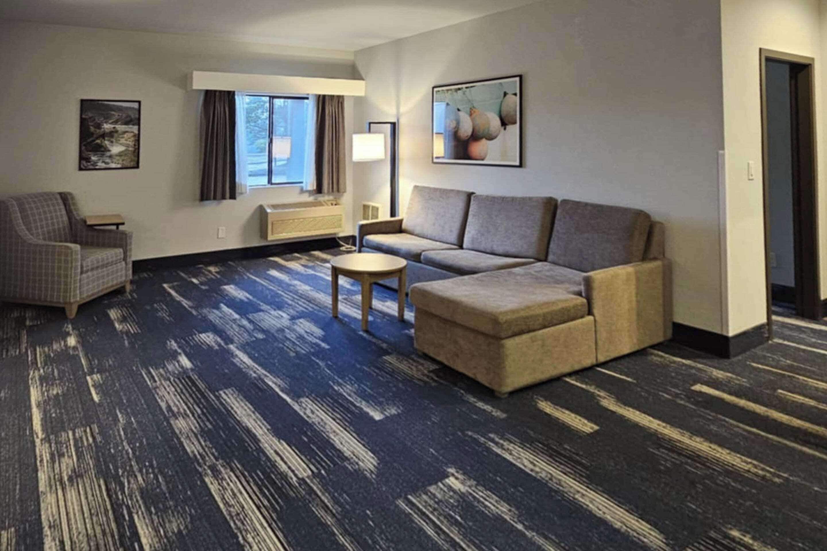 A hotel room with a sectional sofa, a small round table, a chair, and a window with curtains.