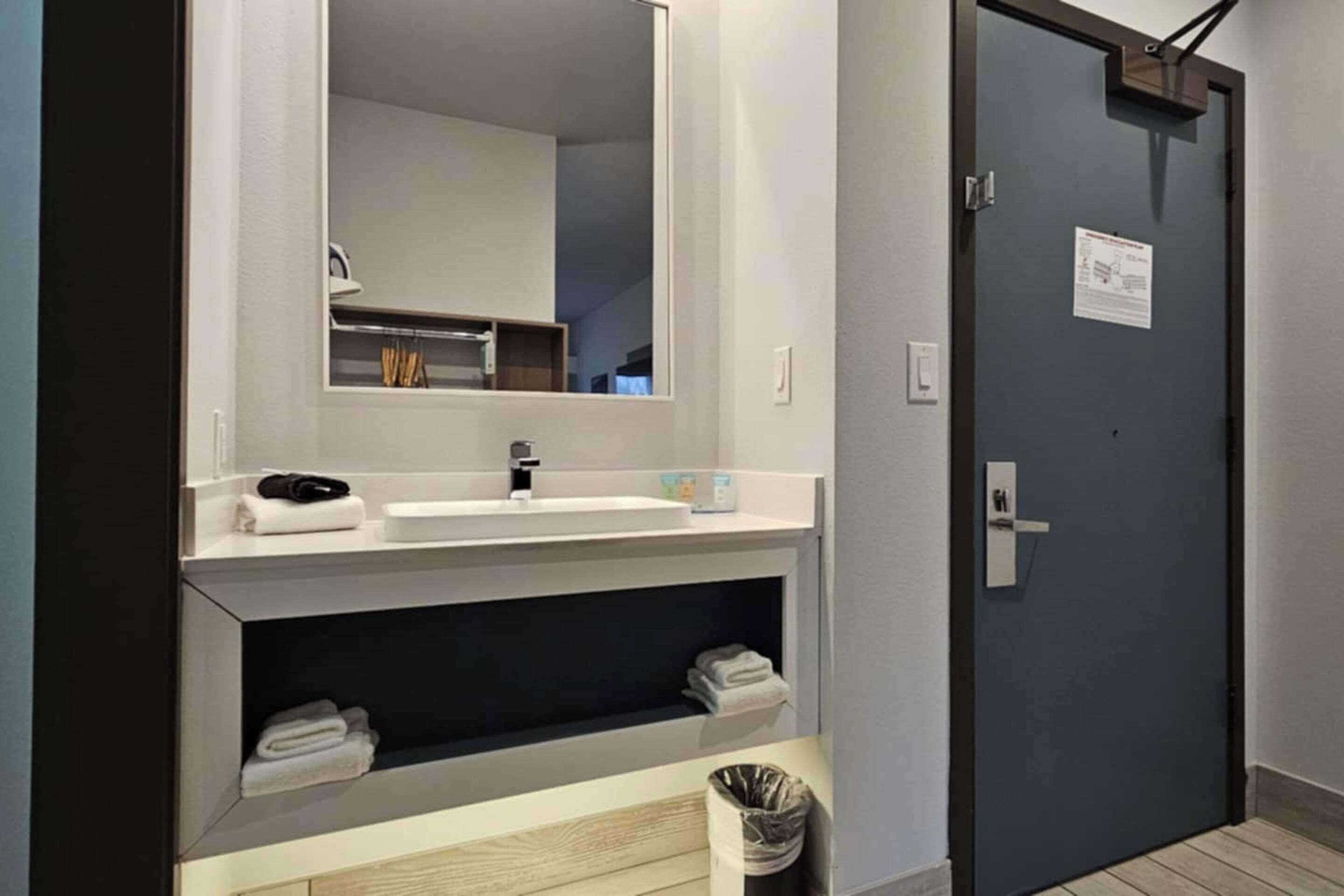 A modern bathroom with a white sink, mirror, and shelves for towels. There is a trash bin and a door with a sign on it.