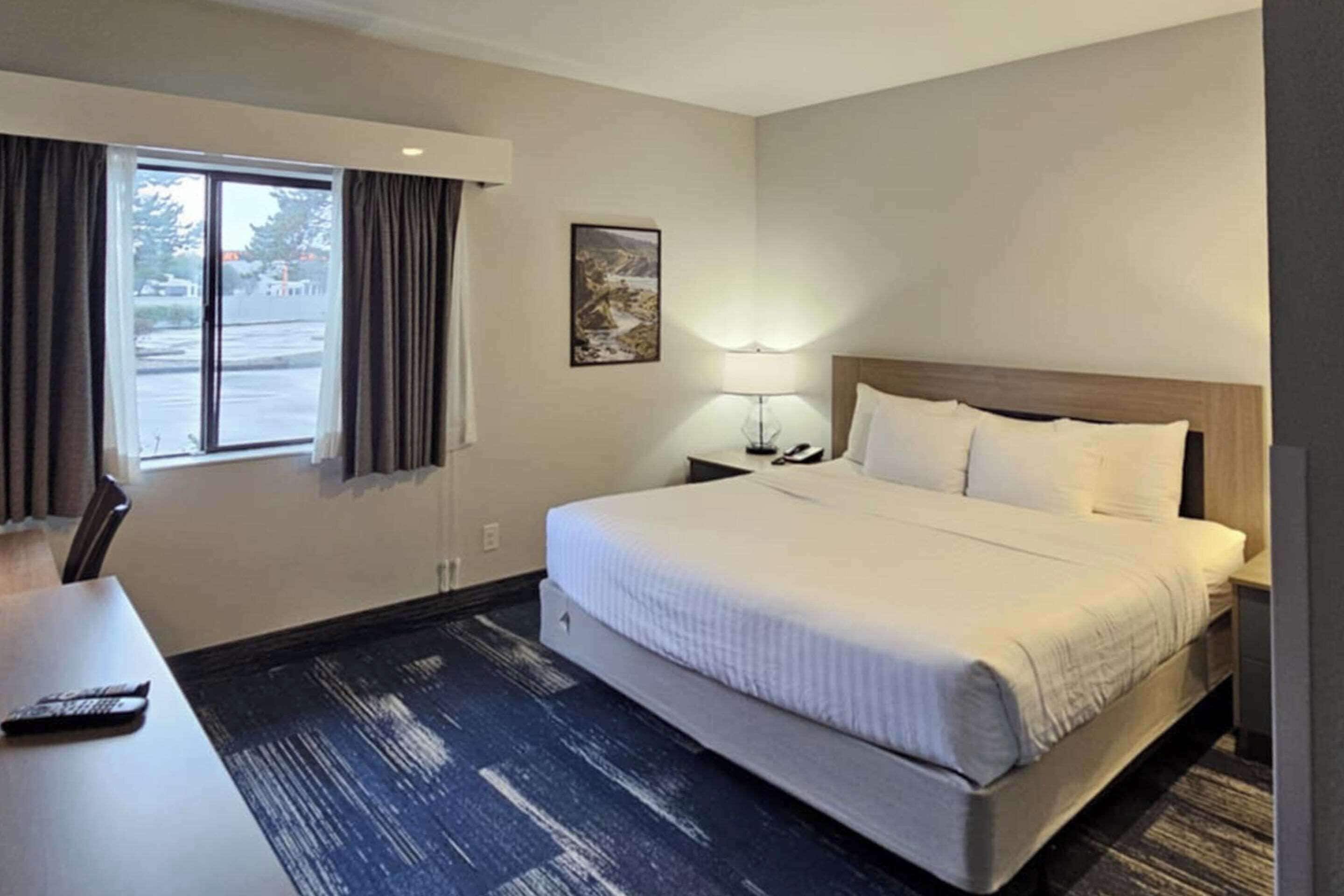A hotel room with a large bed, a desk, a chair, a window with curtains, a lamp, and a painting on the wall.