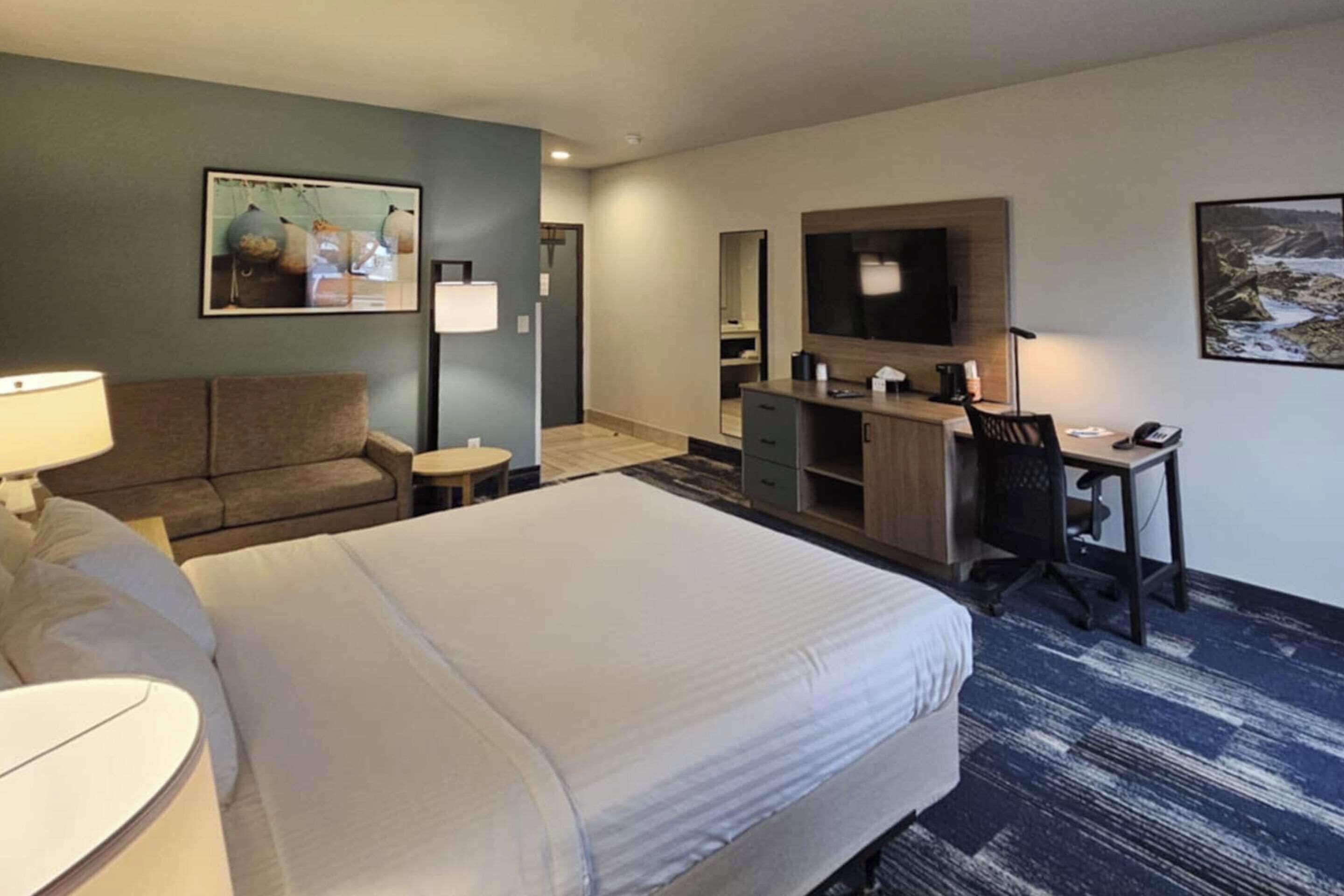 A hotel room with a large bed, a desk with a chair, a sofa, a TV, and a small table with a lamp.