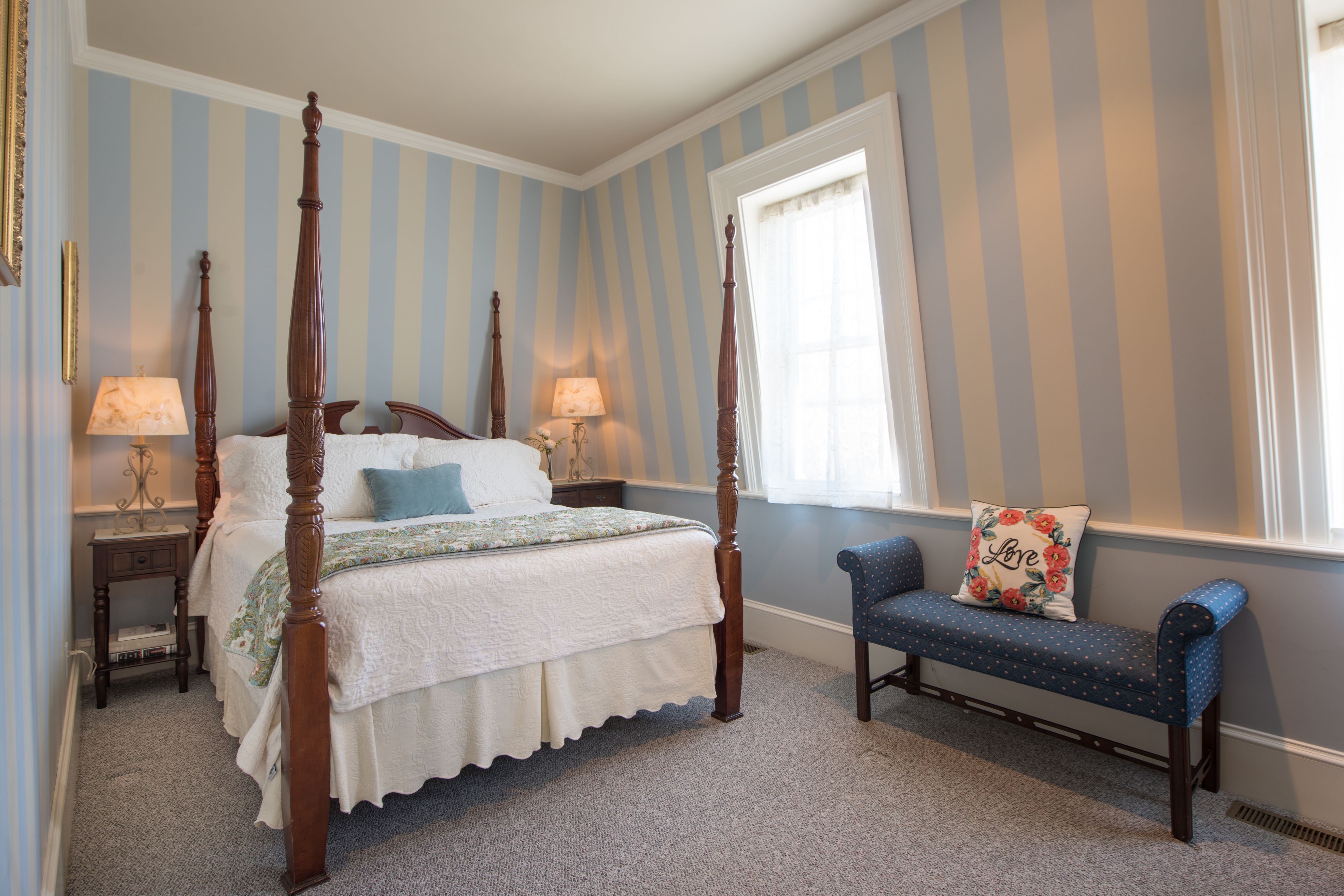 Un lit à baldaquin avec un couvre-lit blanc et des oreillers bleus, une table de chevet en bois avec une lampe, et un banc recouvert d’un tissu bleu agrémenté d’un coussin à motifs floraux.