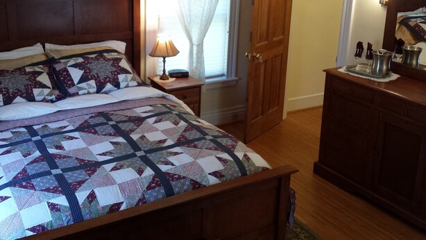 A neatly made bed with a patterned quilt, a bedside lamp, a wooden nightstand, a wooden door, and a dresser with a mirror. - Beautiful Valley Guesthouse (Scottsville)