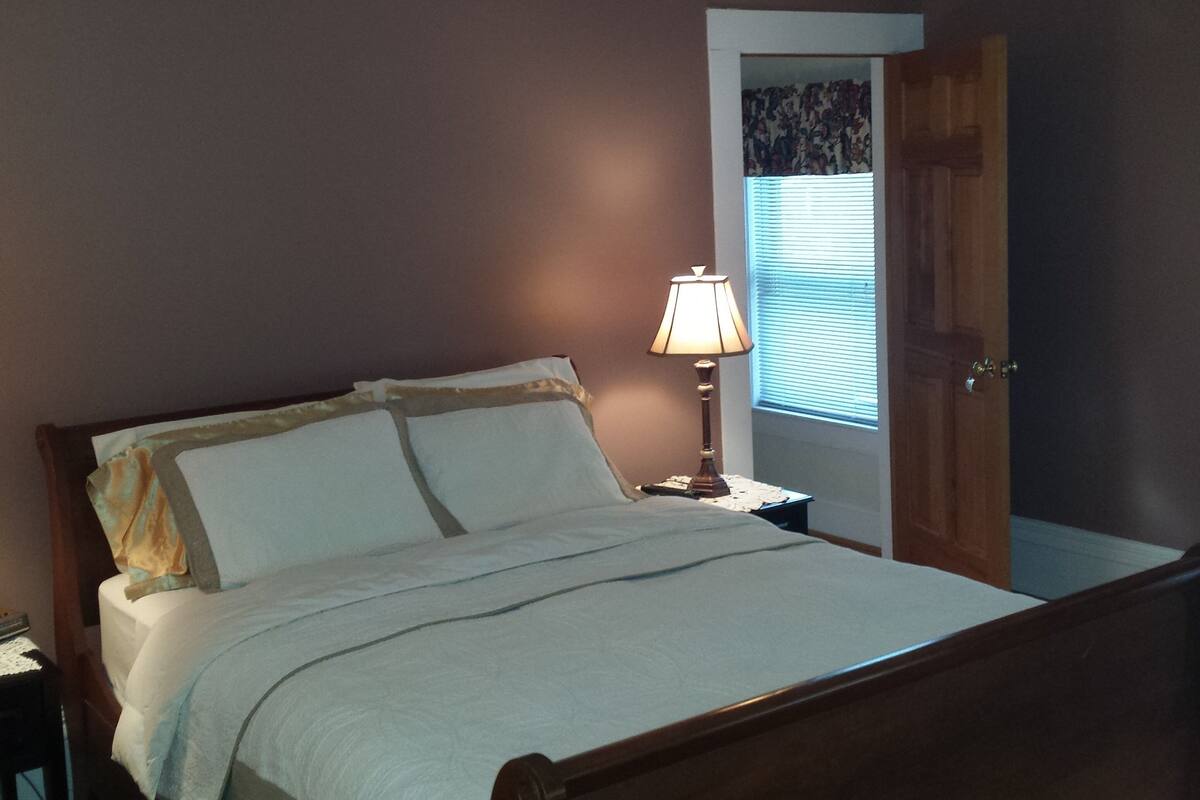 A bedroom with a bed, a bedside table with a lamp, a window with blinds, and a door.