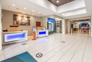 Vestíbulo de hotel con mostradores de recepción, máquina expendedora y zona de estar con televisor.
