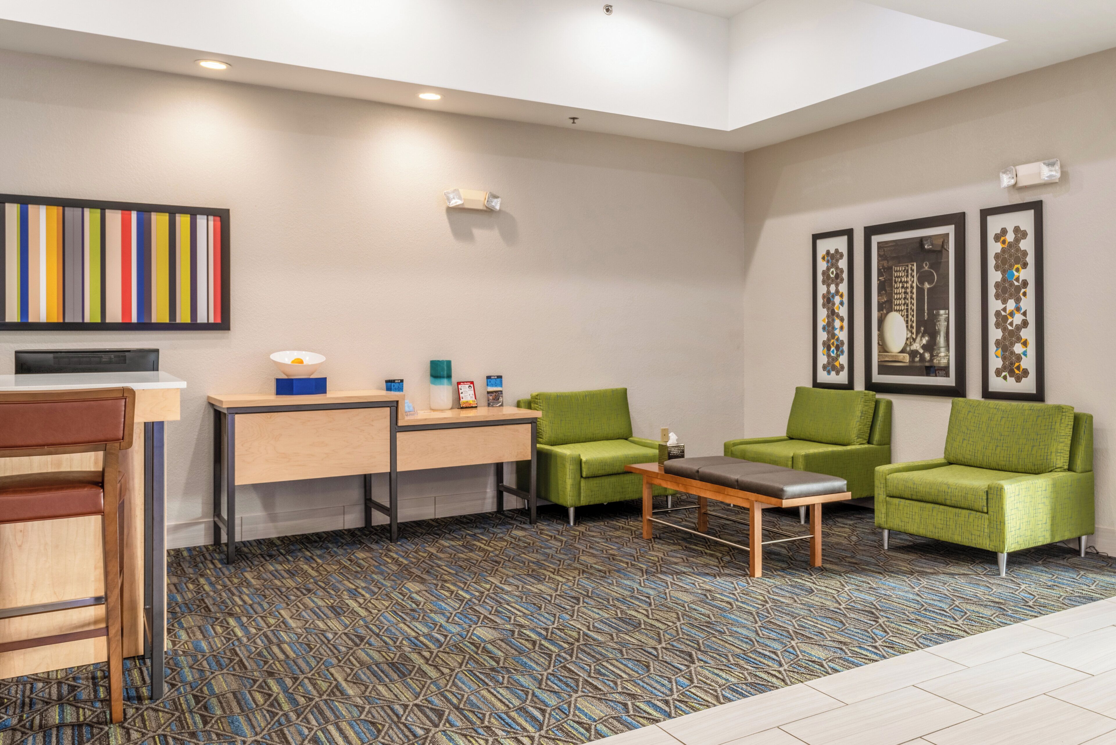 A hotel room with a desk, two green armchairs, a small table, a patterned carpet, and artwork on the walls.
