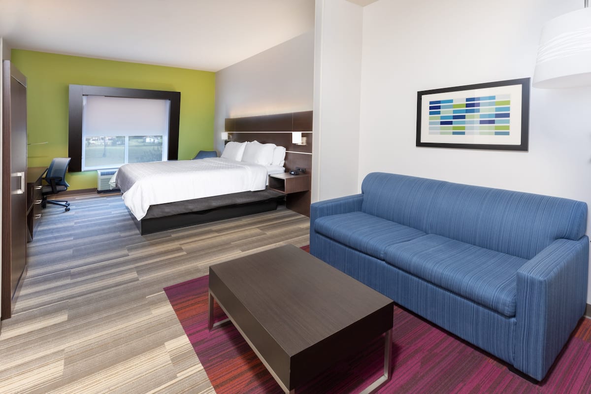 A hotel room with a bed, a blue sofa, a coffee table, a desk with a chair, and a window with blinds.