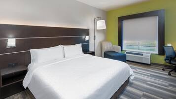 Quarto de hotel com uma cama grande, uma escrivaninha com abajur, uma cadeira e uma janela com vista.
