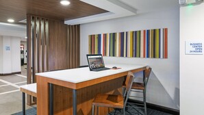 A modern office space with a wooden desk, a laptop, and a chair. A colorful striped wall art is visible, and a sign indicates the business center is open 24 hours. - Holiday Inn Express Picayune-Stennis Space Center by IHG (Picayune)
