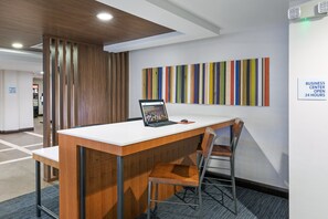A modern office space with a wooden desk, a laptop, and a chair. A colorful striped wall art is visible, and a sign indicates the business center is open 24 hours. - Holiday Inn Express Picayune-Stennis Space Center by IHG (Picayune)