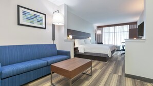 Una habitación de hotel con un sofá azul, una mesa de centro de madera, una cama con ropa blanca y un ventanal con cortinas.