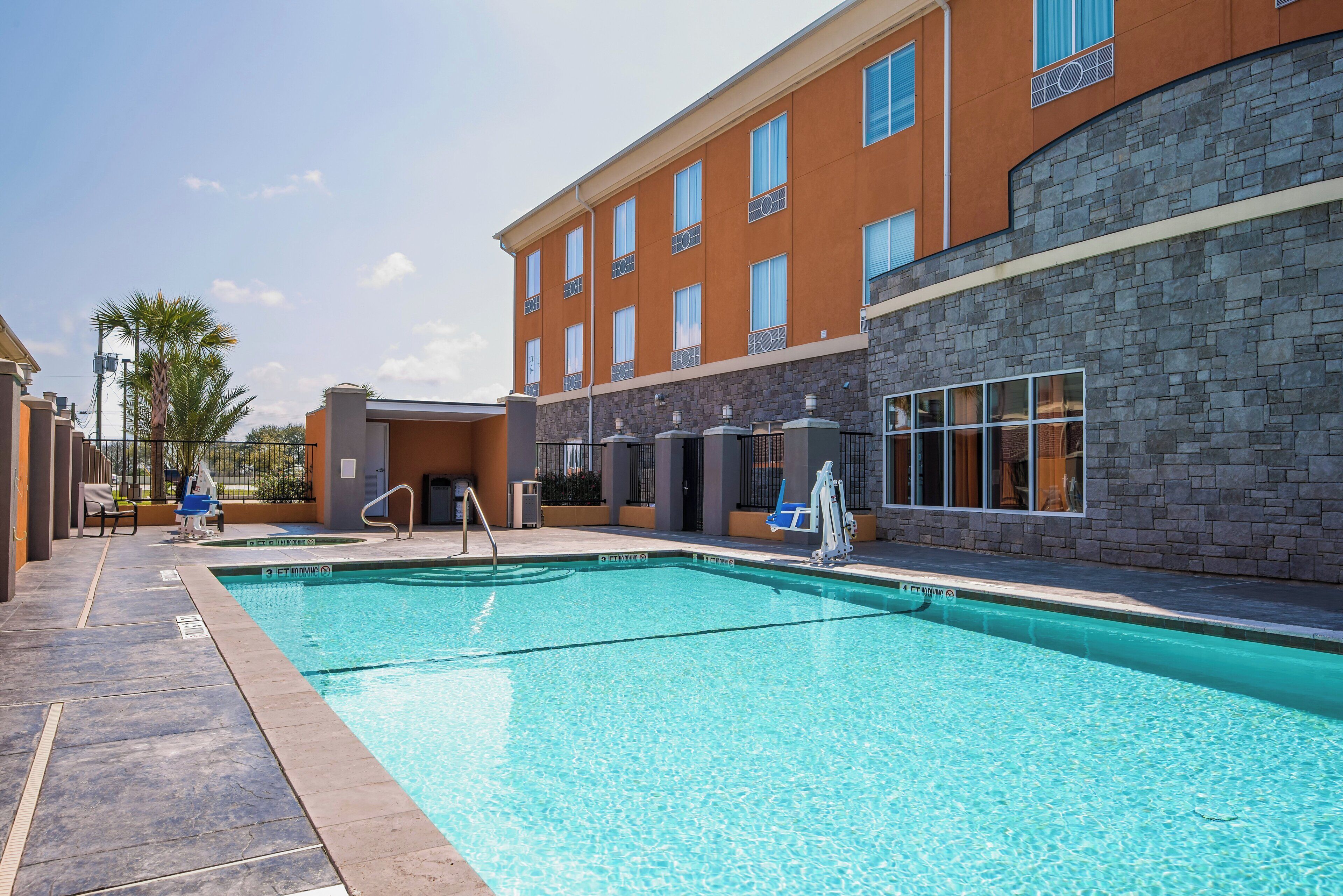 A swimming pool with a clear blue water surface, surrounded by a stone wall and a building with multiple windows.