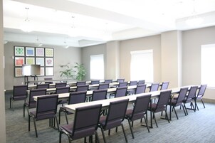 Un salón de conferencias con filas de sillas y mesas, una pantalla de proyector y una pared con cuadros enmarcados.