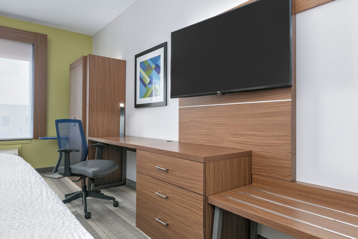 A hotel room with a wooden desk, a chair, a bed, a TV, and a framed picture on the wall.