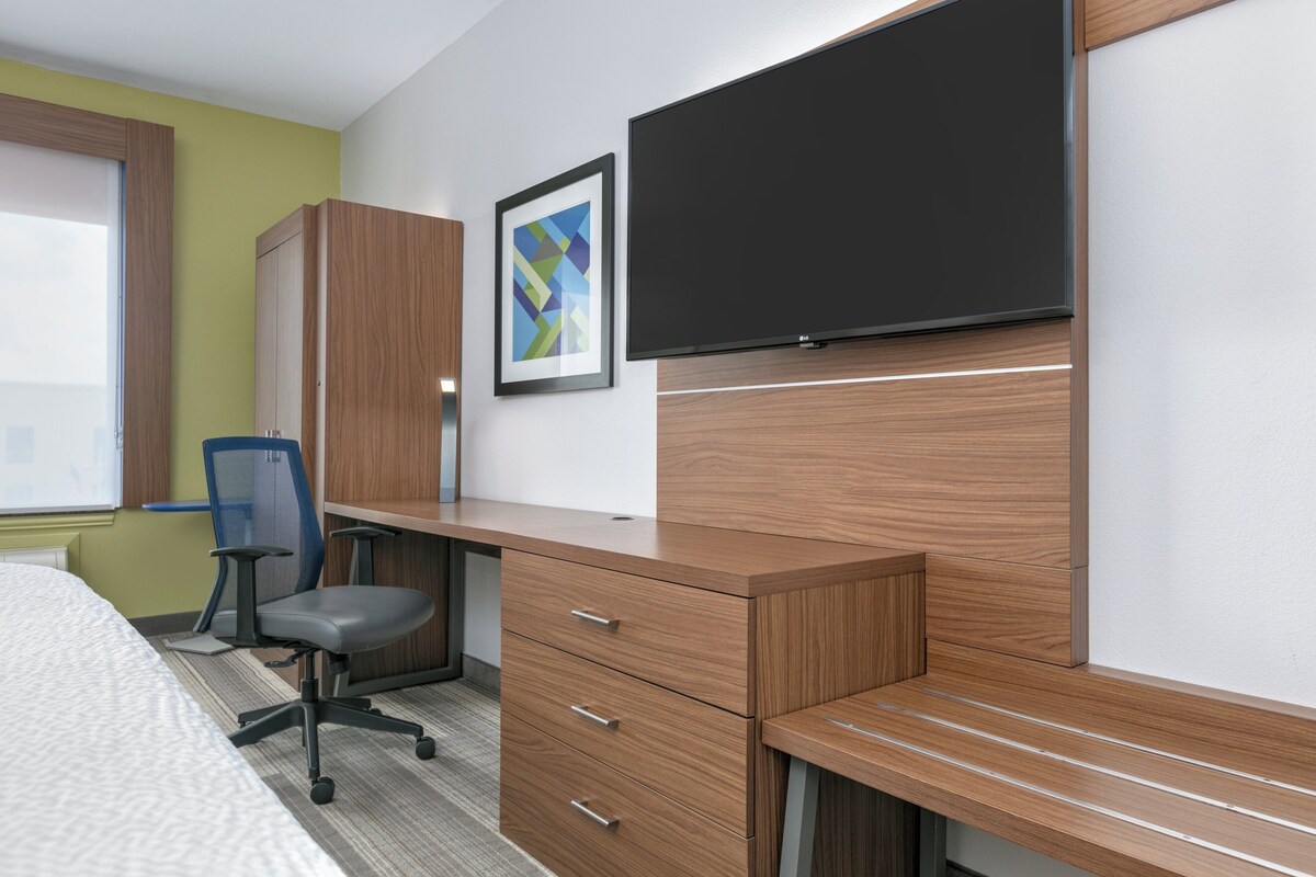 A hotel room with a wooden desk, a chair, a bed, a TV, and a framed picture on the wall.