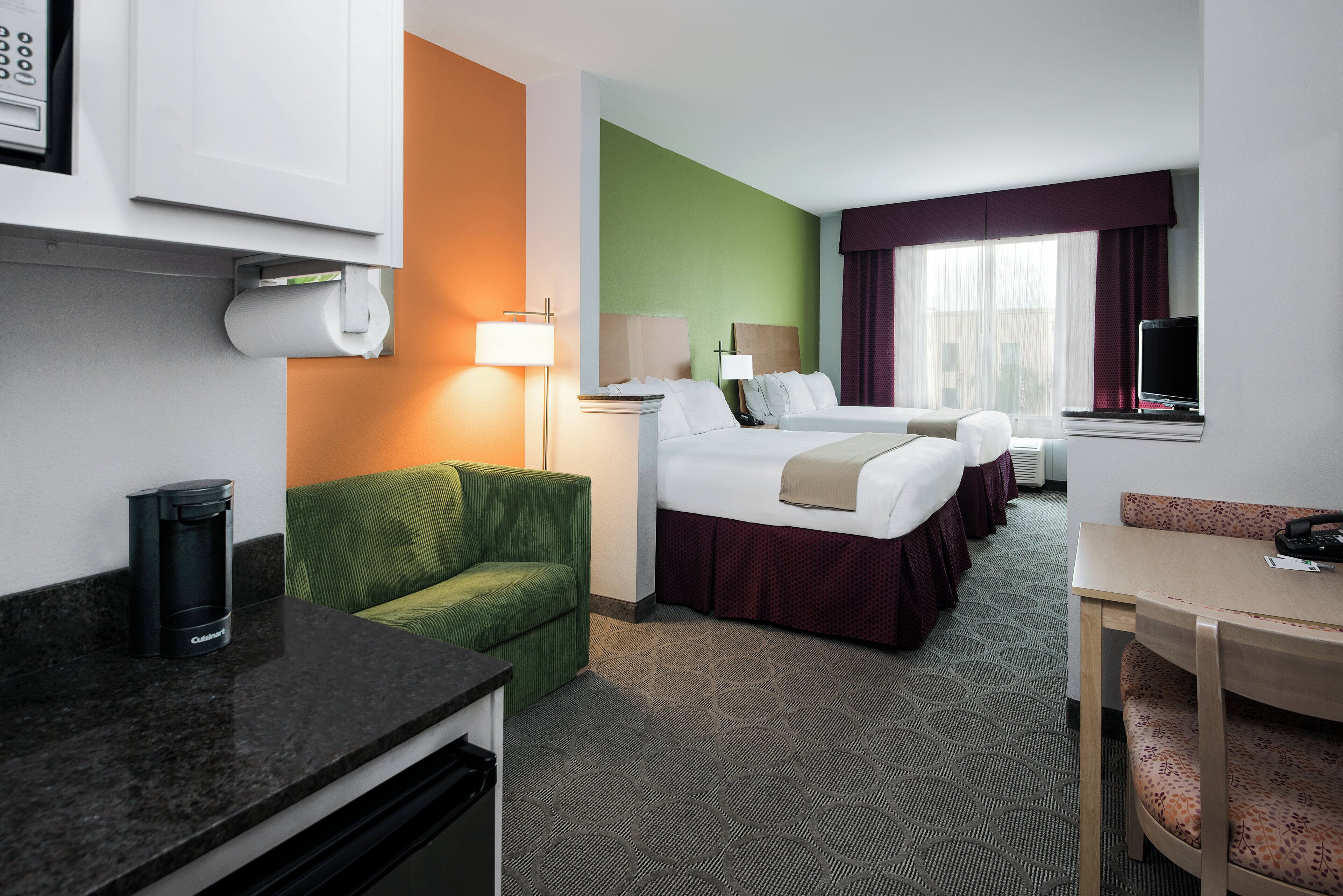 A hotel room with a bed, a desk, a chair, a sofa, a trash bin, and a window with curtains.