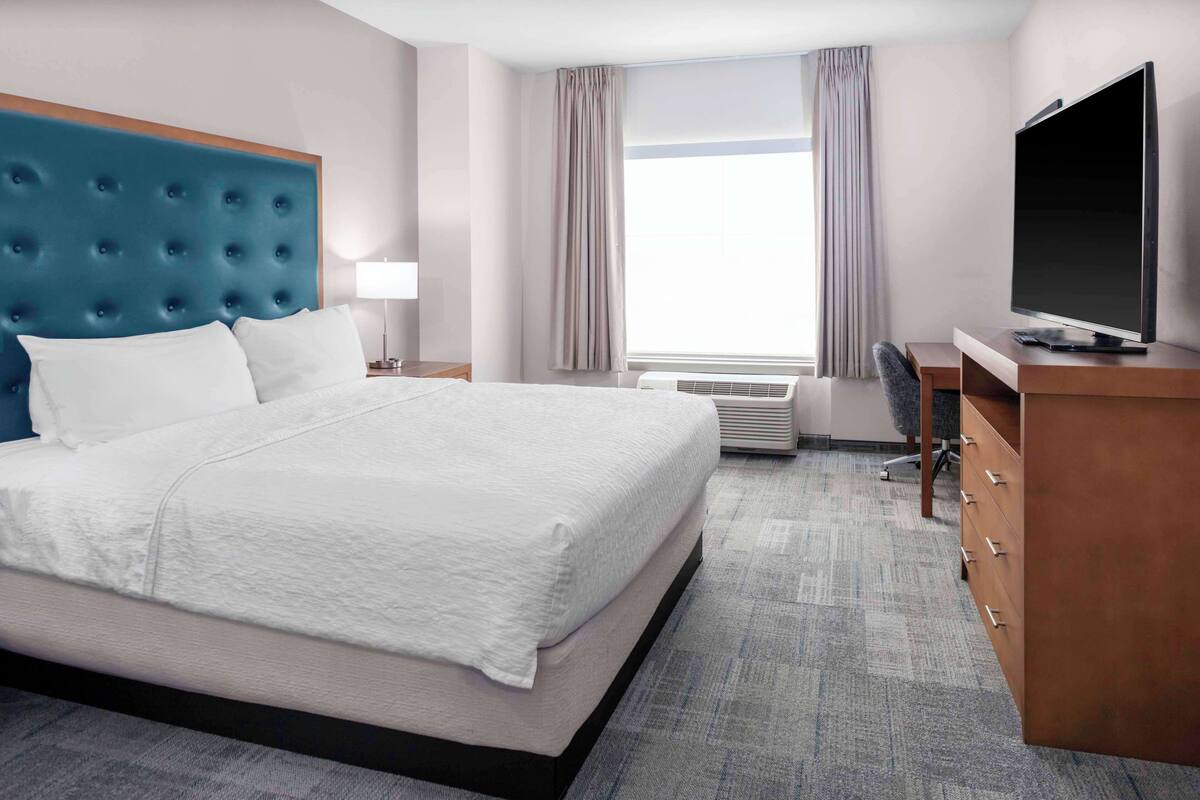 A hotel room with a large bed, a desk with a flat-screen TV, a chair, a lamp, and a window with curtains.