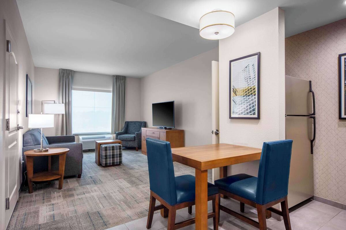 A modern hotel room with a dining area, a sofa, a television, and a refrigerator.