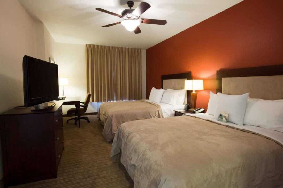 A hotel room with two beds, a desk with a television, a ceiling fan, and a window with curtains.