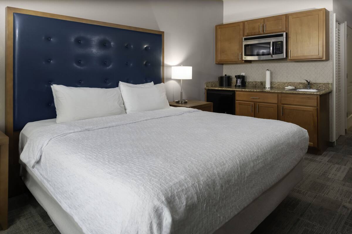 A hotel room with a large bed, a headboard, a nightstand, a lamp, a microwave, and a kitchenette.