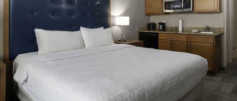 A hotel room with a large bed, a headboard, a nightstand, a lamp, a microwave, and a kitchenette.