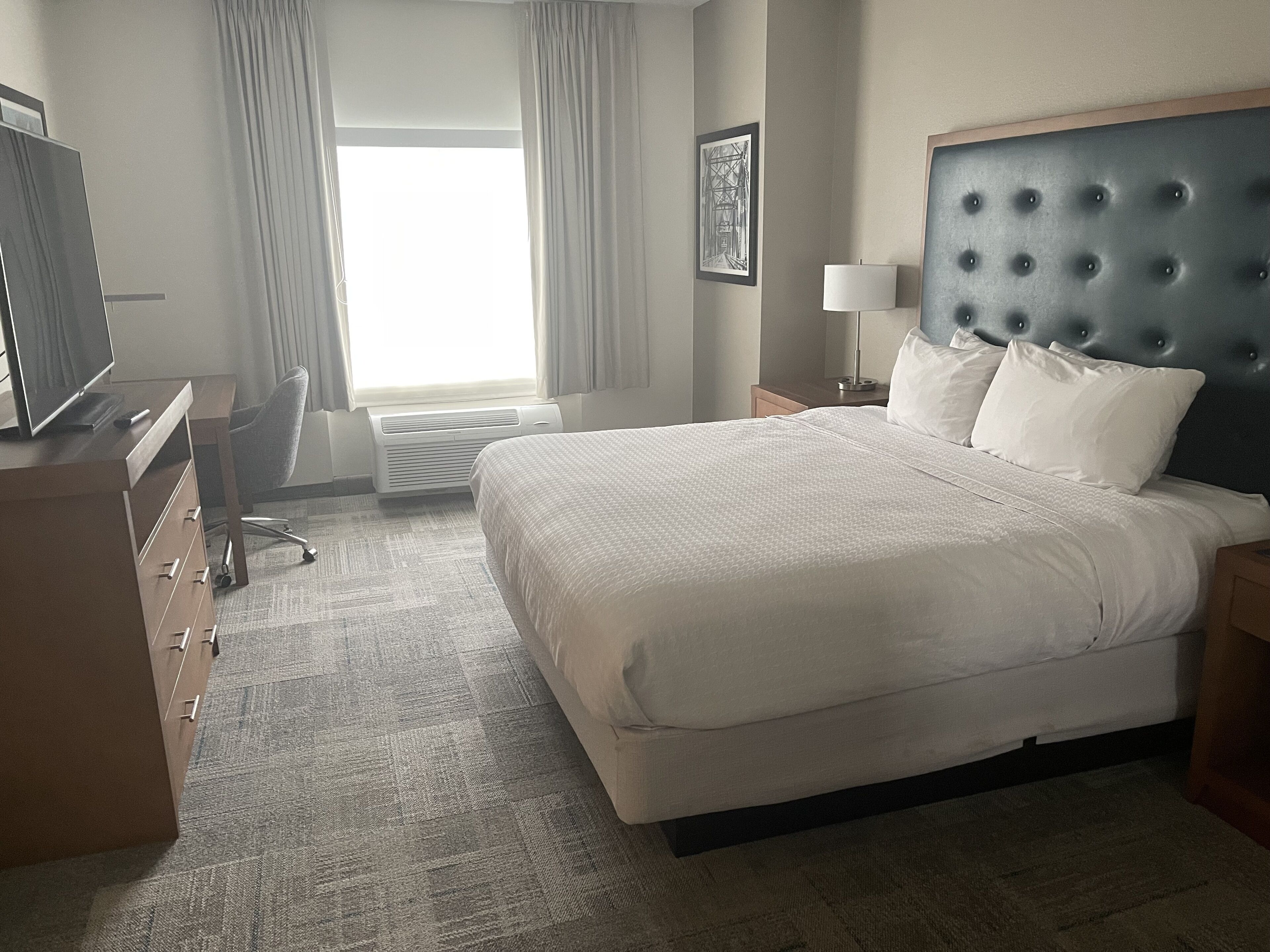 A hotel room with a large bed, a desk with a computer, a chair, a lamp, and a framed picture on the wall.