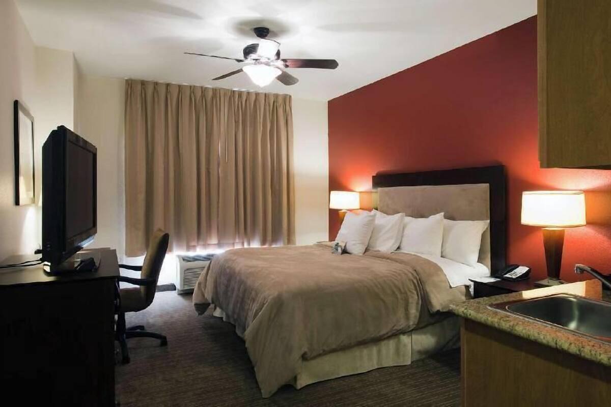 A hotel room with a bed, a desk with a computer, a chair, a ceiling fan, a window with curtains, and a nightstand with a lamp.